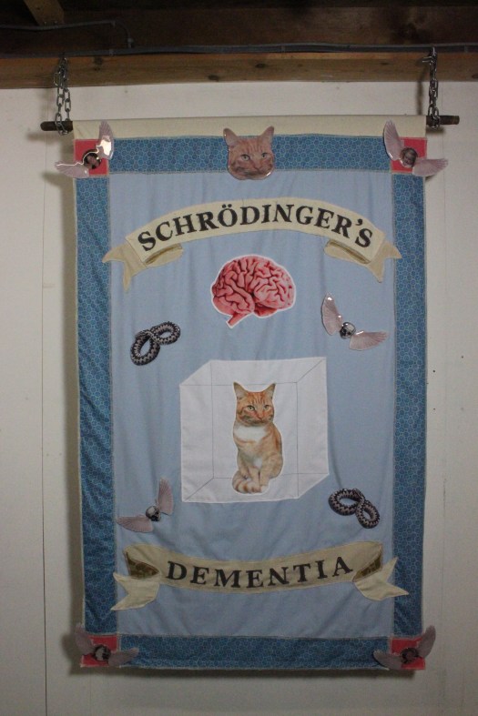 fabric stitched banner with two pennants that have the words 'Schrödinger’s Dementia' and glass badges with memento mori imagery: a snake and a winged skull. Plus a fabric printed cat stitched onto a febric cube